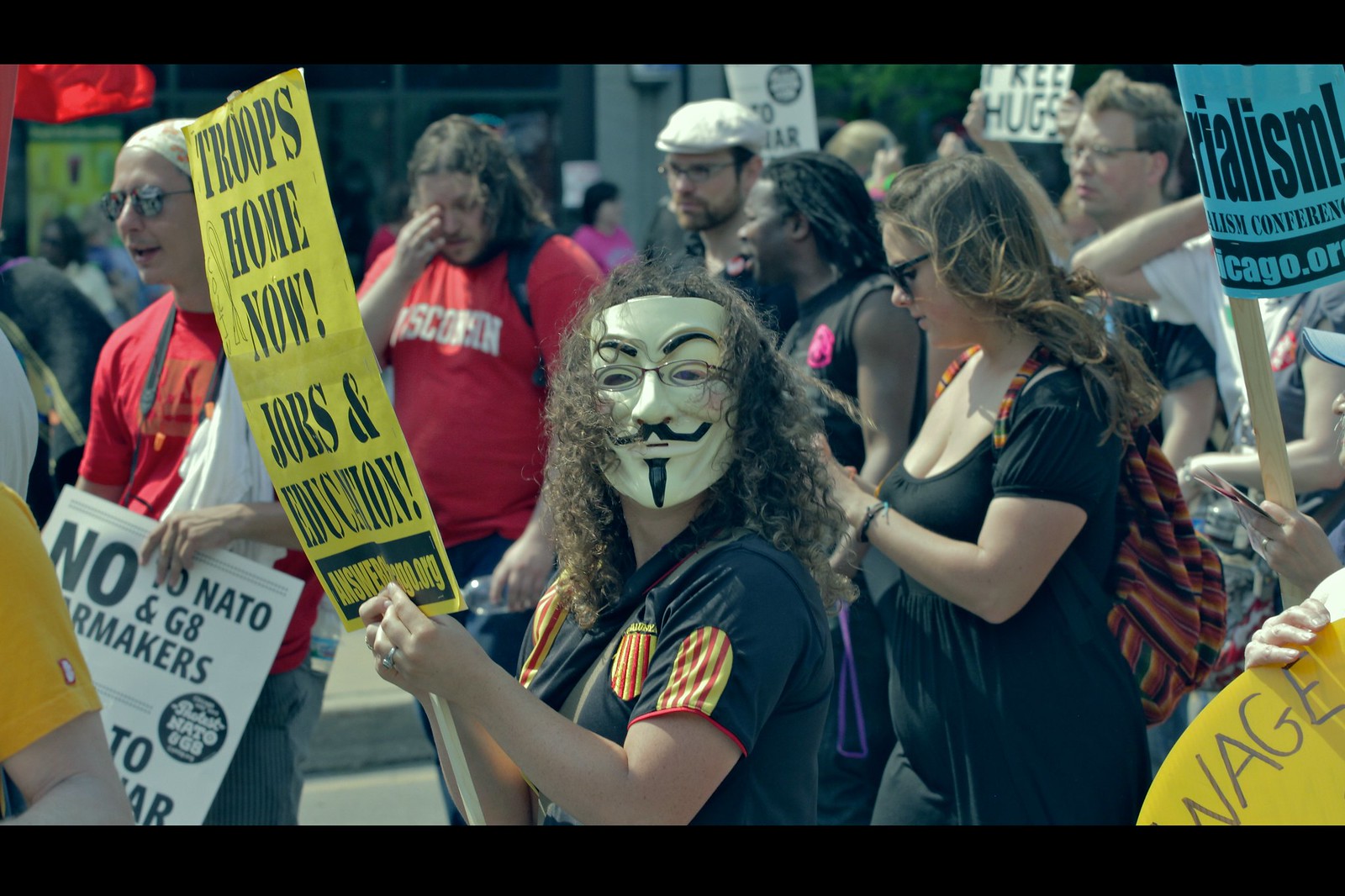 Protestor wearing a Guy Fawkes mask holds a yellow sign that says "TROOPS HOME NOW! JOBS & EDUCATION!" during a crowded political rally. Chaos, conviction, and questionable font choices ensue.